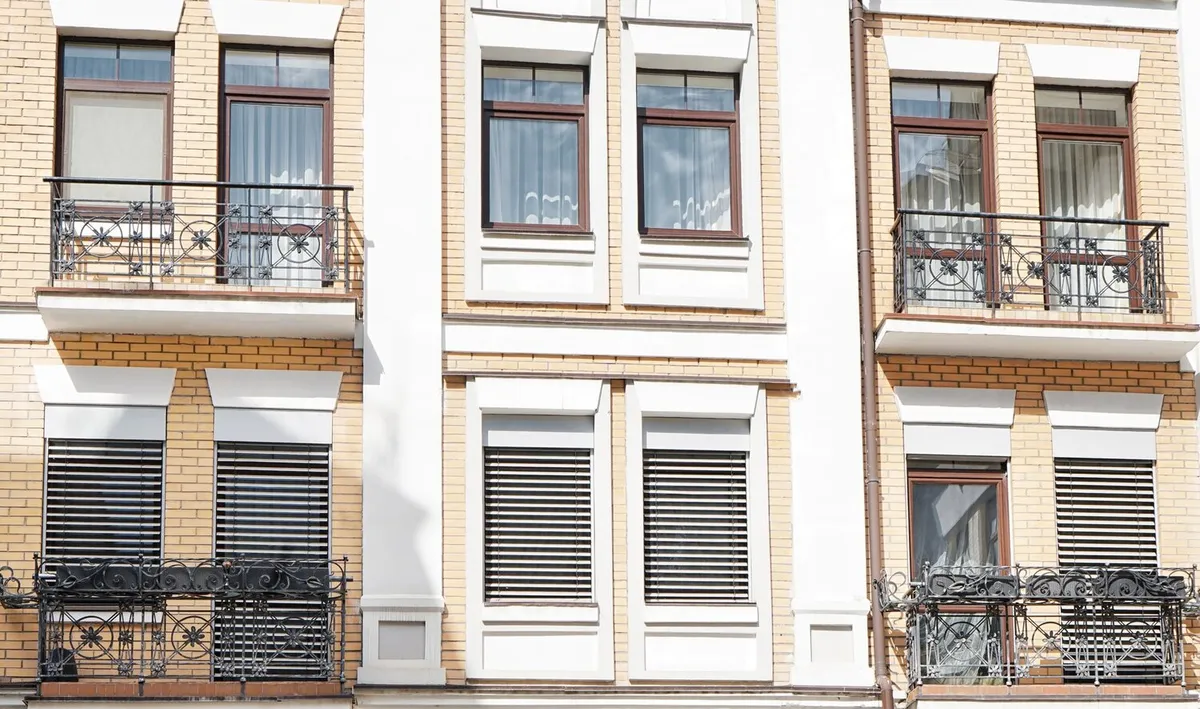 Photo d'illustration : façade d'immeuble en brique claire avec balcons en fer forgé, volets roulants baissés aux fenêtres du bas